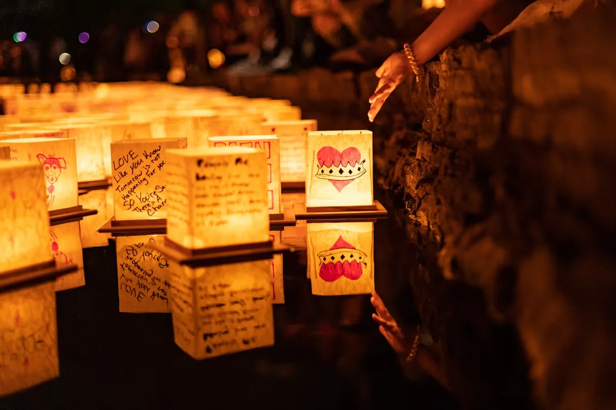 Example photography of people beside decorated lanterns floating on water at night; not this event. Location and season unverified.
